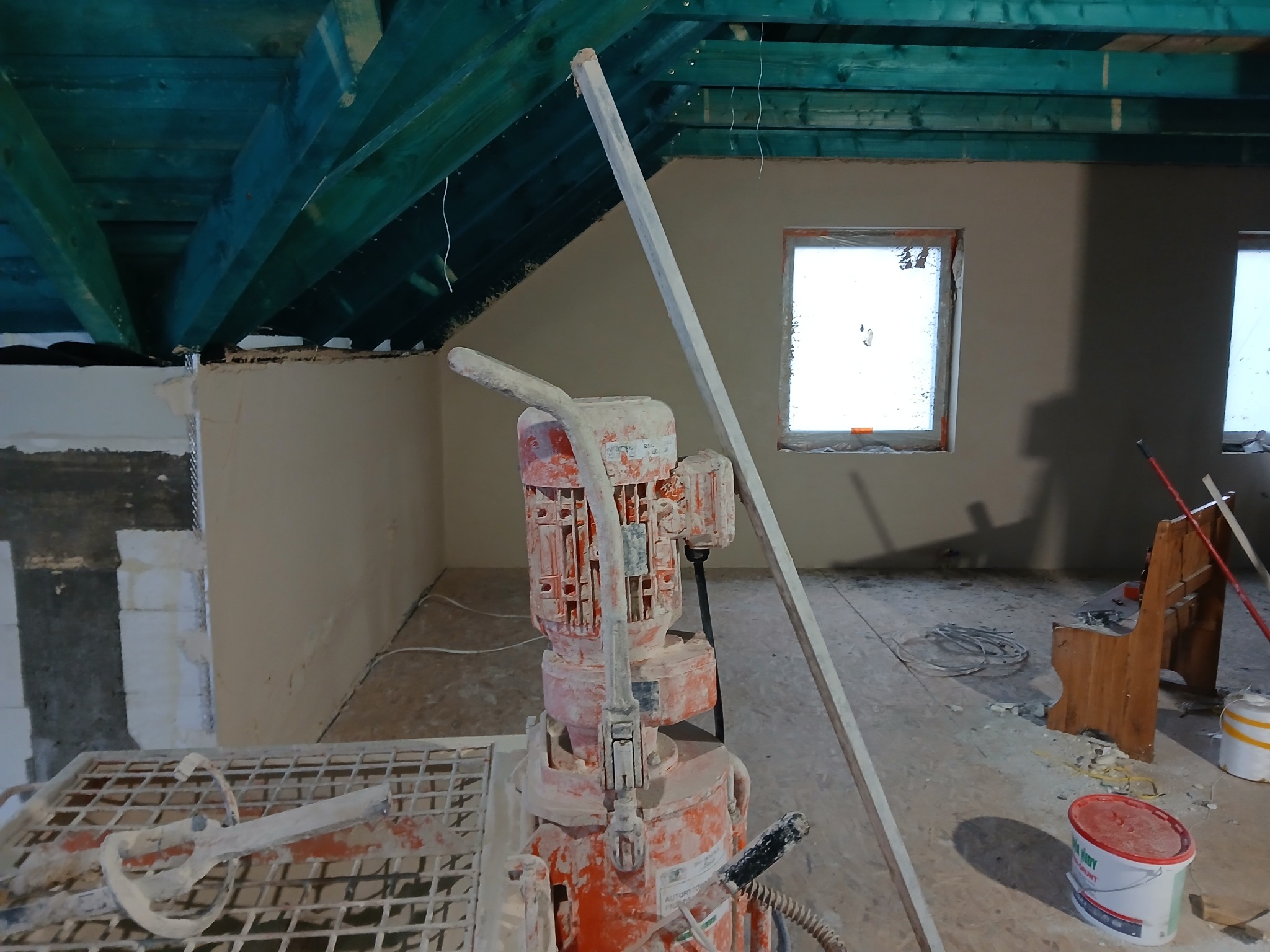 Wnętrze w trakcie prac tynkarskich. Widoczna maszyna do tynkowania, ściany pokryte świeżym tynkiem oraz drewniana konstrukcja dachu. Dwa okna w tle.