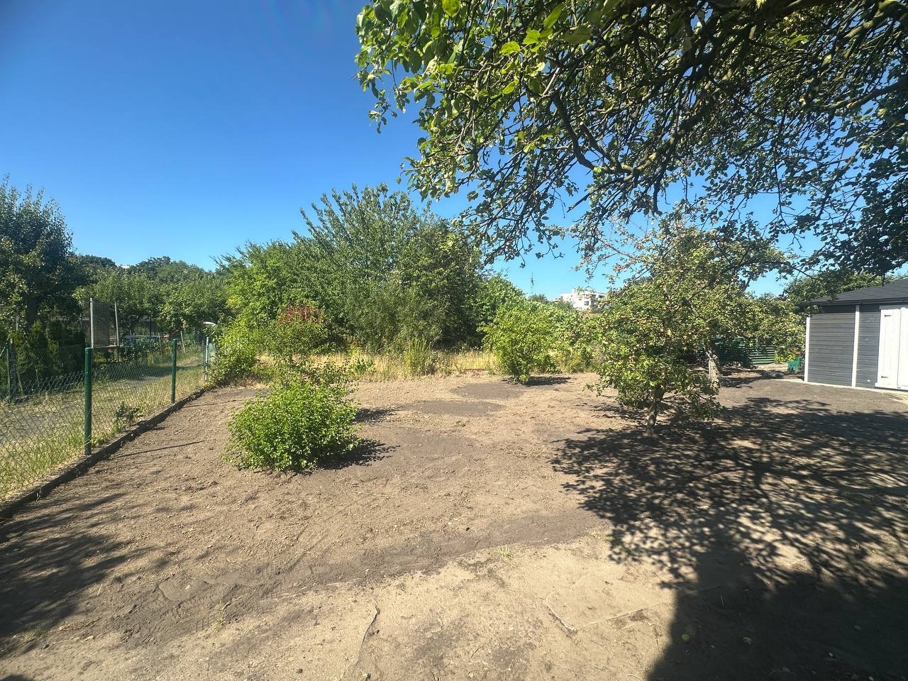 Przygotowany teren pod przyszły ogród, z widocznymi śladami po usuniętych roślinach, ogrodzeniem i drzewami w tle, w słoneczny dzień.