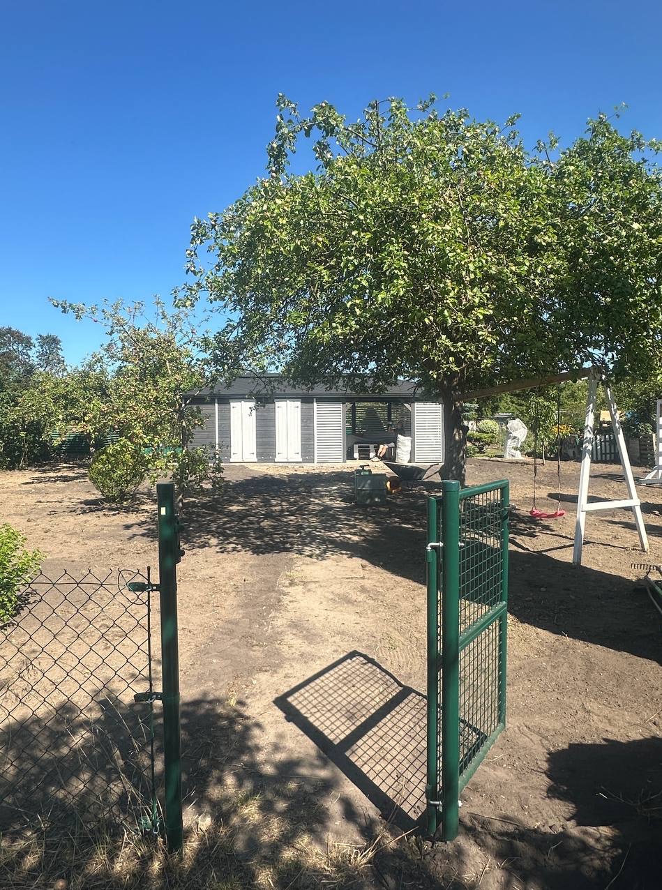 Ogród z otwartą, zieloną furtką, prowadzącą do szarej altany z białymi okiennicami, pod rozłożystym drzewem w słoneczny dzień. Widoczny plac zabaw i narzędzia.