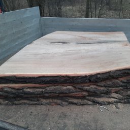 ForestPro - Stos pociętych, surowych desek z korą, ułożonych na pace samochodu dostawczego. Widoczne ślady cięcia i kora na krawędziach. W tle drzewa bez liści.