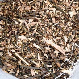 ForestPro - Worek wypełniony świeżo pociętymi gałęziami i drobnymi kawałkami drewna, leżący na trawie. Widoczny fragment buta osoby stojącej obok.