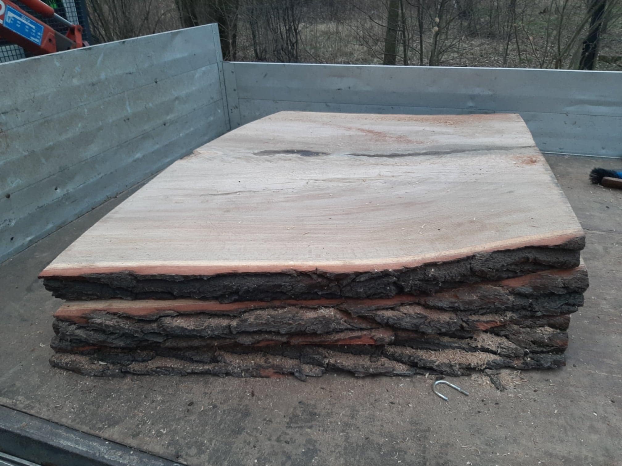 Stos pociętych, surowych desek z korą, ułożonych na pace samochodu dostawczego. Widoczne ślady cięcia i kora na krawędziach. W tle drzewa bez liści.