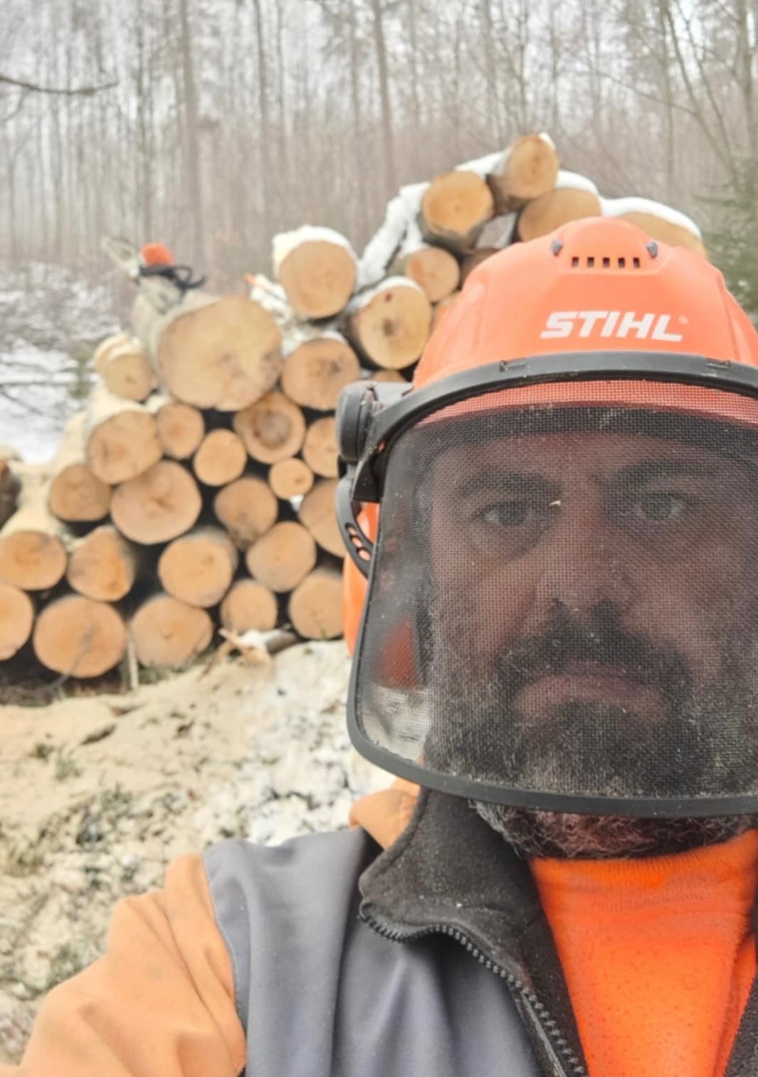 Mężczyzna w kasku Stihl z siatką ochronną na tle stosu ściętych pni drzew w zimowym lesie. Widoczne ślady śniegu i piły spalinowej.