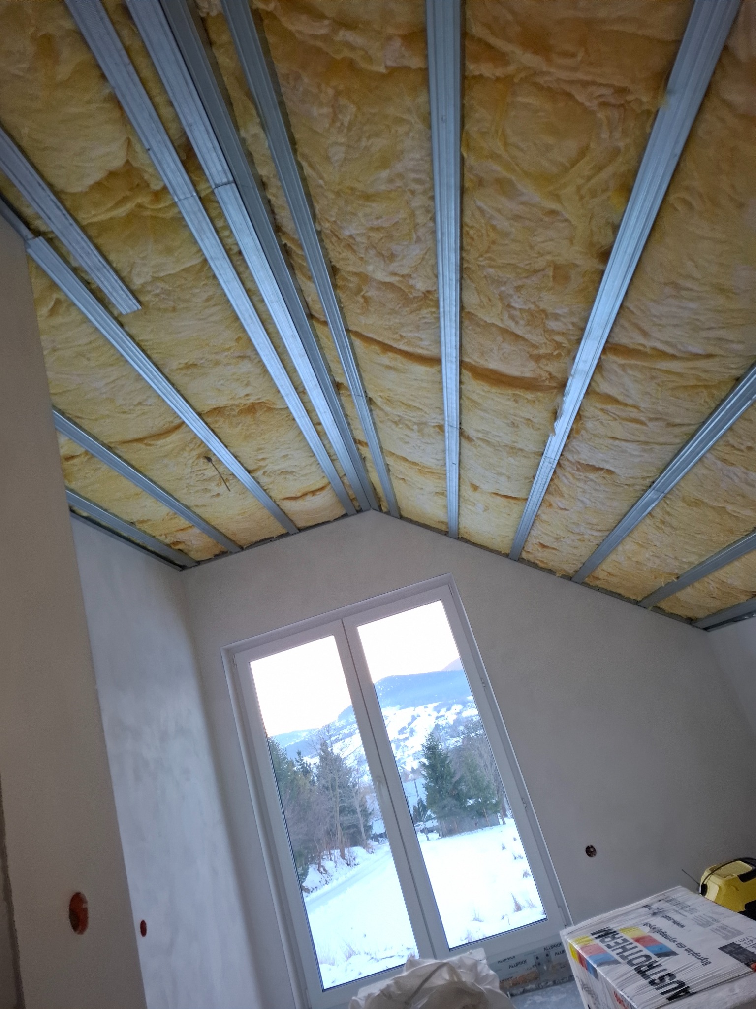 Sufit podwieszany z izolacją z wełny mineralnej i metalową konstrukcją, widok z dołu, okno z widokiem na zimowy krajobraz, etap prac wykończeniowych.