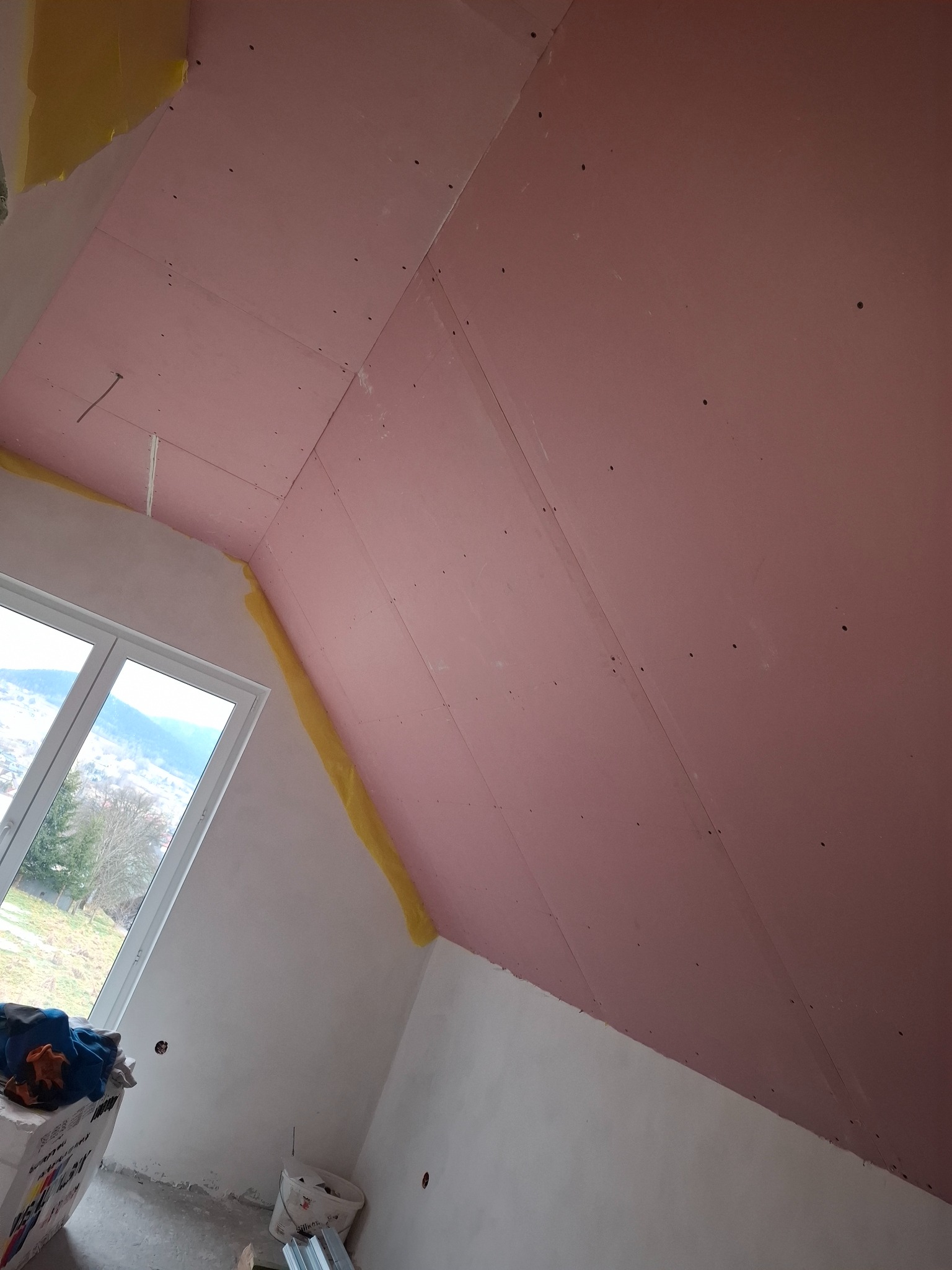 Poddasze z zamontowaną płytą karton-gips w kolorze różowym, widoczne łączenia płyt i wkręty. Okno w tle z widokiem na zewnątrz, fragmenty żółtej taśmy.