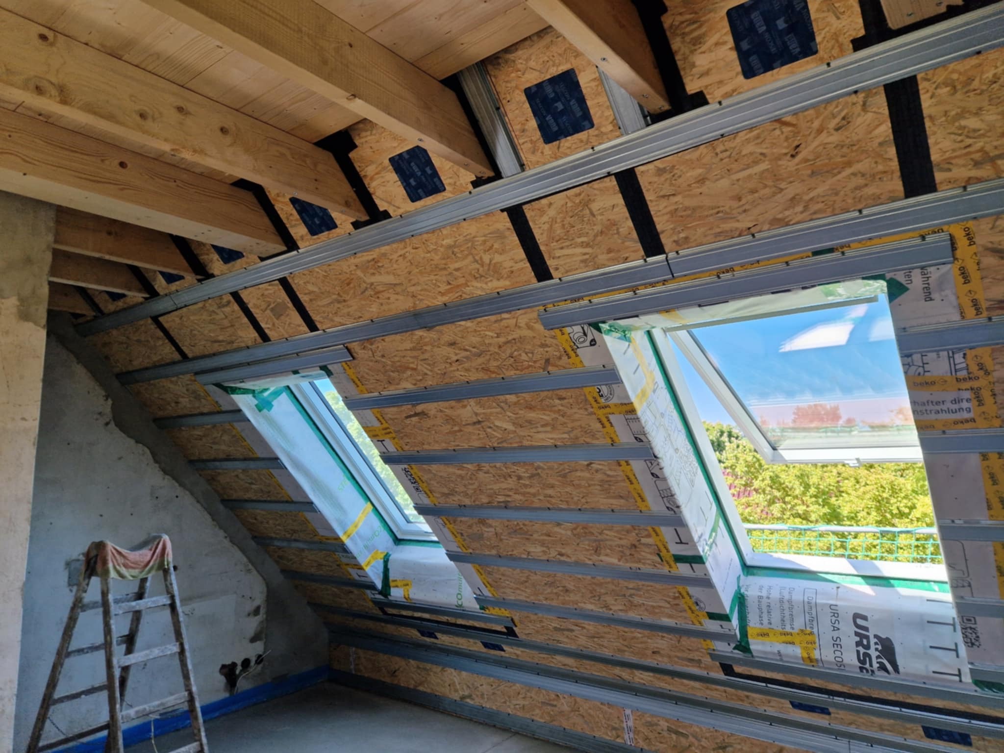 Montaż płyt OSB na poddaszu z oknami dachowymi, widoczne metalowe profile i drewniana konstrukcja, drabina oparta o surową ścianę. Prace wykończeniowe w trakcie.