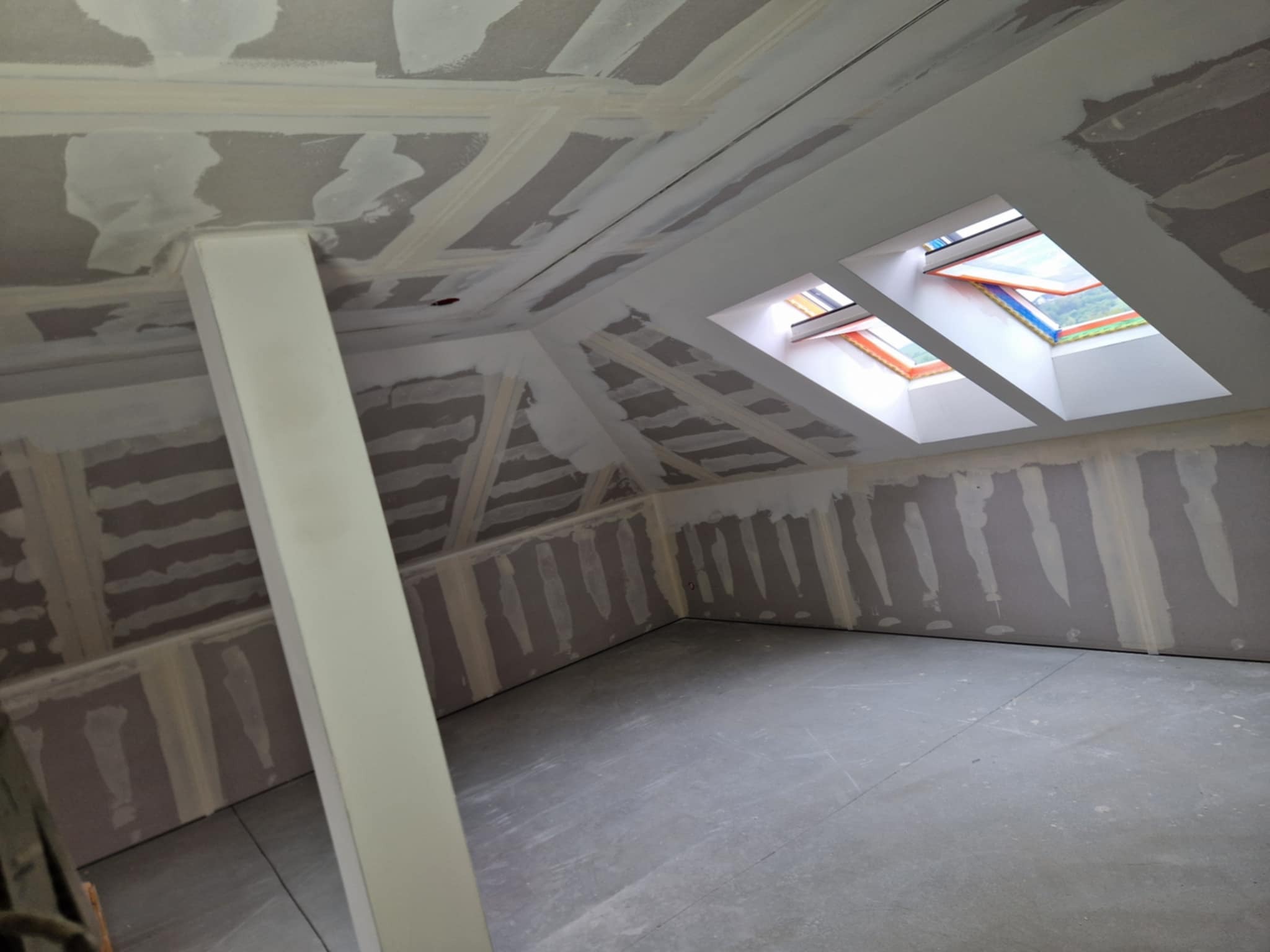 Pokój na poddaszu w trakcie wykończenia: ściany z płyt karton-gips, szpachlowane łączenia, widoczne okna dachowe i podpora konstrukcyjna.