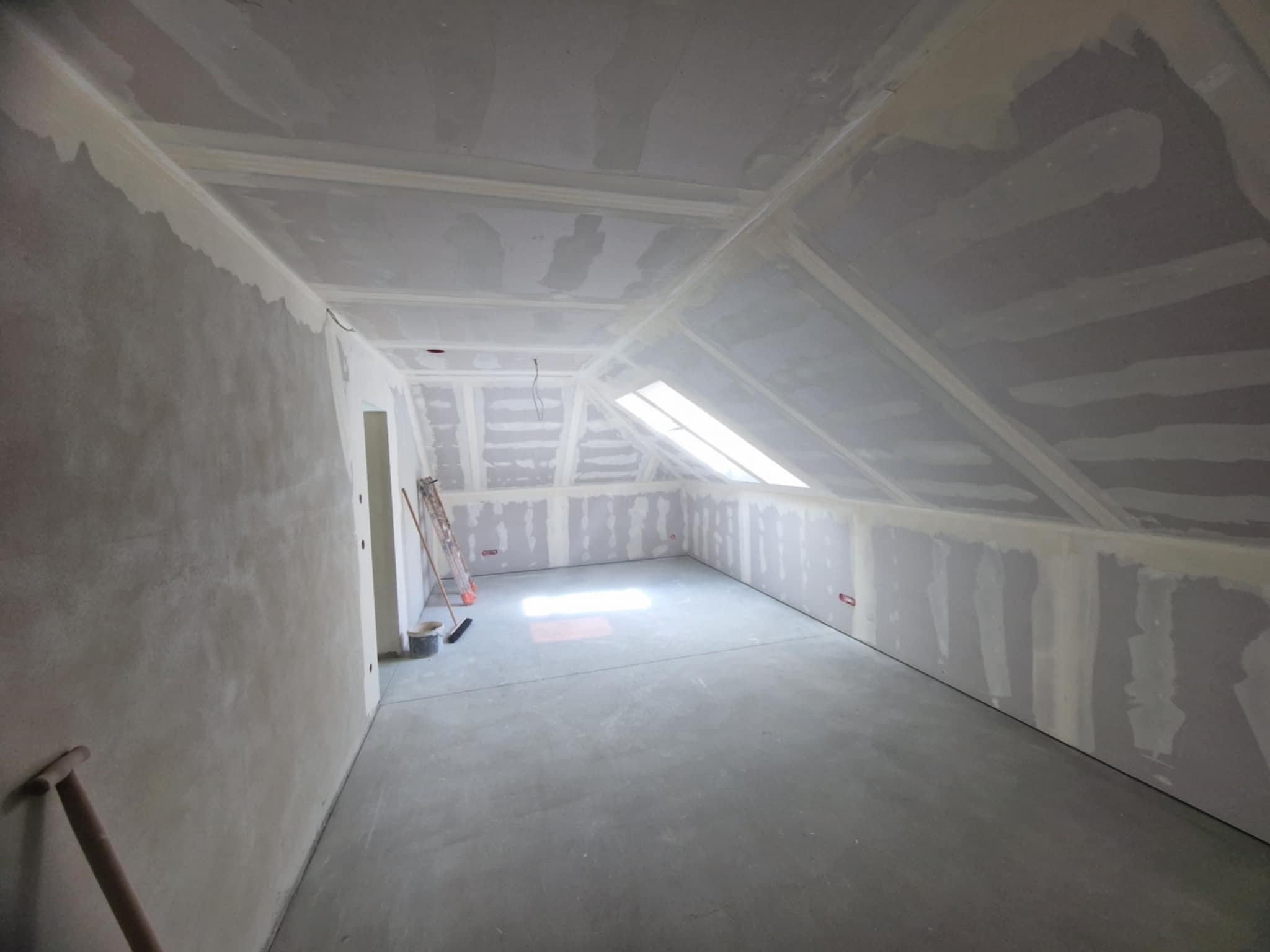 Poddasze po szpachlowaniu i montażu płyt karton-gips, widoczne łączenia płyt, narzędzia malarskie, okno dachowe. Ściany przygotowane do malowania.