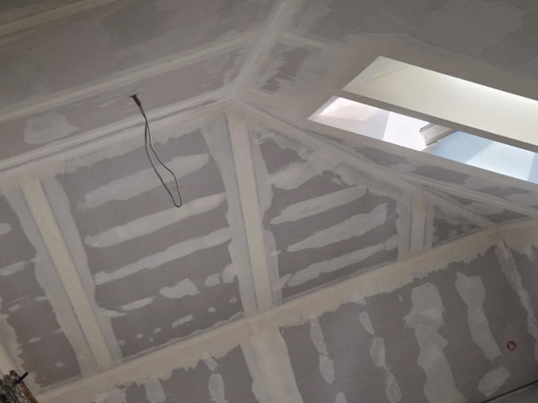 Skośne poddasze z oknem dachowym, wykończone płytami kartonowo-gipsowymi i wstępnie wyszpachlowane, widoczne łączenia płyt. Wystający kabel elektryczny.