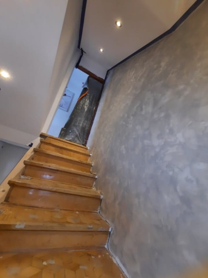 Klatka schodowa w trakcie remontu: drewniane schody, ściana z efektem dekoracyjnym zabezpieczona taśmą malarską. Widoczne oświetlenie punktowe.