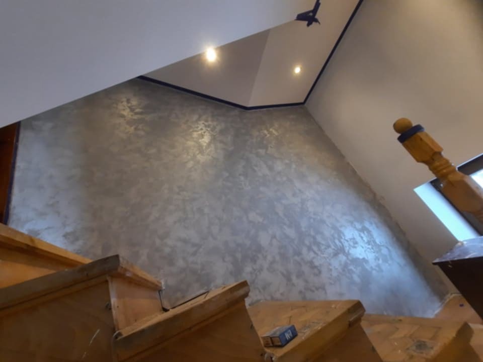 Dekoracyjna ściana w odcieniach szarości z efektem metalicznego połysku, widoczna z perspektywy schodów. Na suficie widoczne oświetlenie punktowe i dekoracja.