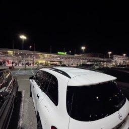 EKOTRANS-GK SPÓŁKA Z OGRANICZONĄ ODPOWIEDZIALNOŚCIĄ - Biały Mercedes GLB 200d na parkingu lotniska nocą, widok z tyłu, oświetlony terminal w tle. Tablice rejestracyjne zamazane.