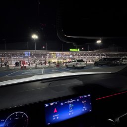 EKOTRANS-GK SPÓŁKA Z OGRANICZONĄ ODPOWIEDZIALNOŚCIĄ - Widok z wnętrza samochodu na parking przed Terminalem C lotniska nocą. Na desce rozdzielczej włączony ekran nawigacji. Parkujące samochody i oświetlony budynek w tle.