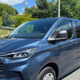 EKOTRANS-GK SPÓŁKA Z OGRANICZONĄ ODPOWIEDZIALNOŚCIĄ - Niebieski Ford Transit Custom z reflektorem LED na tle zieleni i błękitnego nieba. Widoczny fragment tablicy rejestracyjnej.