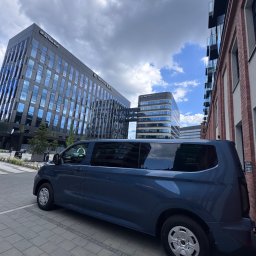 EKOTRANS-GK SPÓŁKA Z OGRANICZONĄ ODPOWIEDZIALNOŚCIĄ - Niebieski bus na tle nowoczesnych biurowców z przeszklonymi fasadami i ceglanego budynku. Kadr z dołu, widoczne chmury.