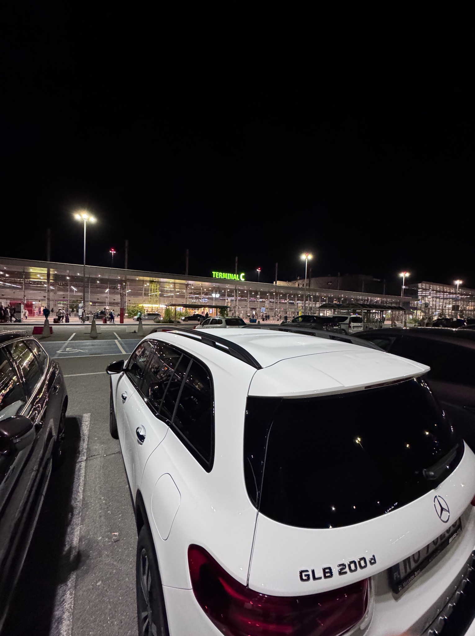 Biały Mercedes GLB 200d na parkingu lotniska nocą, widok z tyłu, oświetlony terminal w tle. Tablice rejestracyjne zamazane.