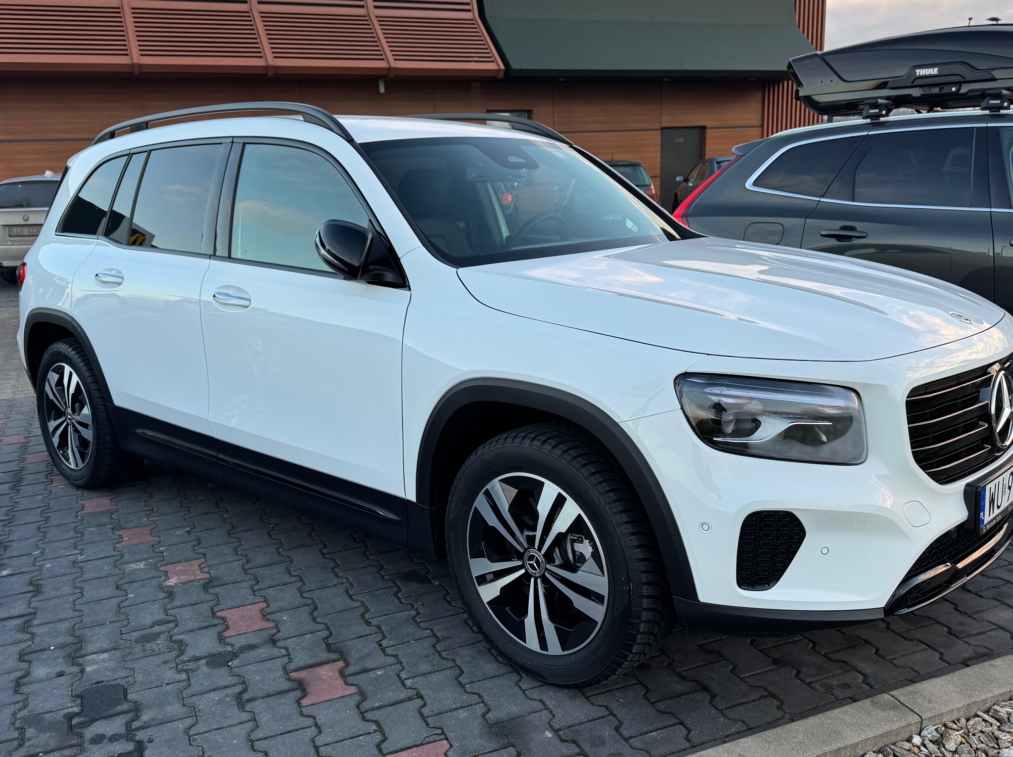 Biały SUV Mercedes-Benz GLB z relingami dachowymi, zaparkowany na kostce brukowej przed budynkiem z drewnianą fasadą. Widoczny fragment drugiego samochodu z bagażnikiem dachowym.