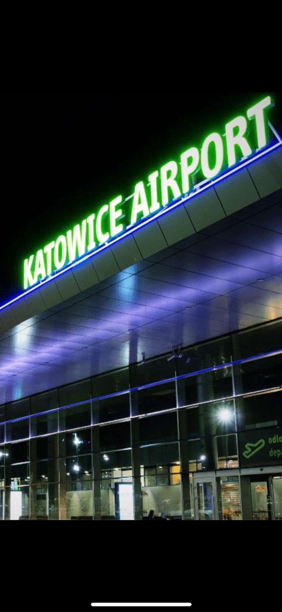 Nocny widok na budynek lotniska Katowice z neonowym napisem. Szklana fasada odbija światła, tworząc nowoczesny, architektoniczny krajobraz.