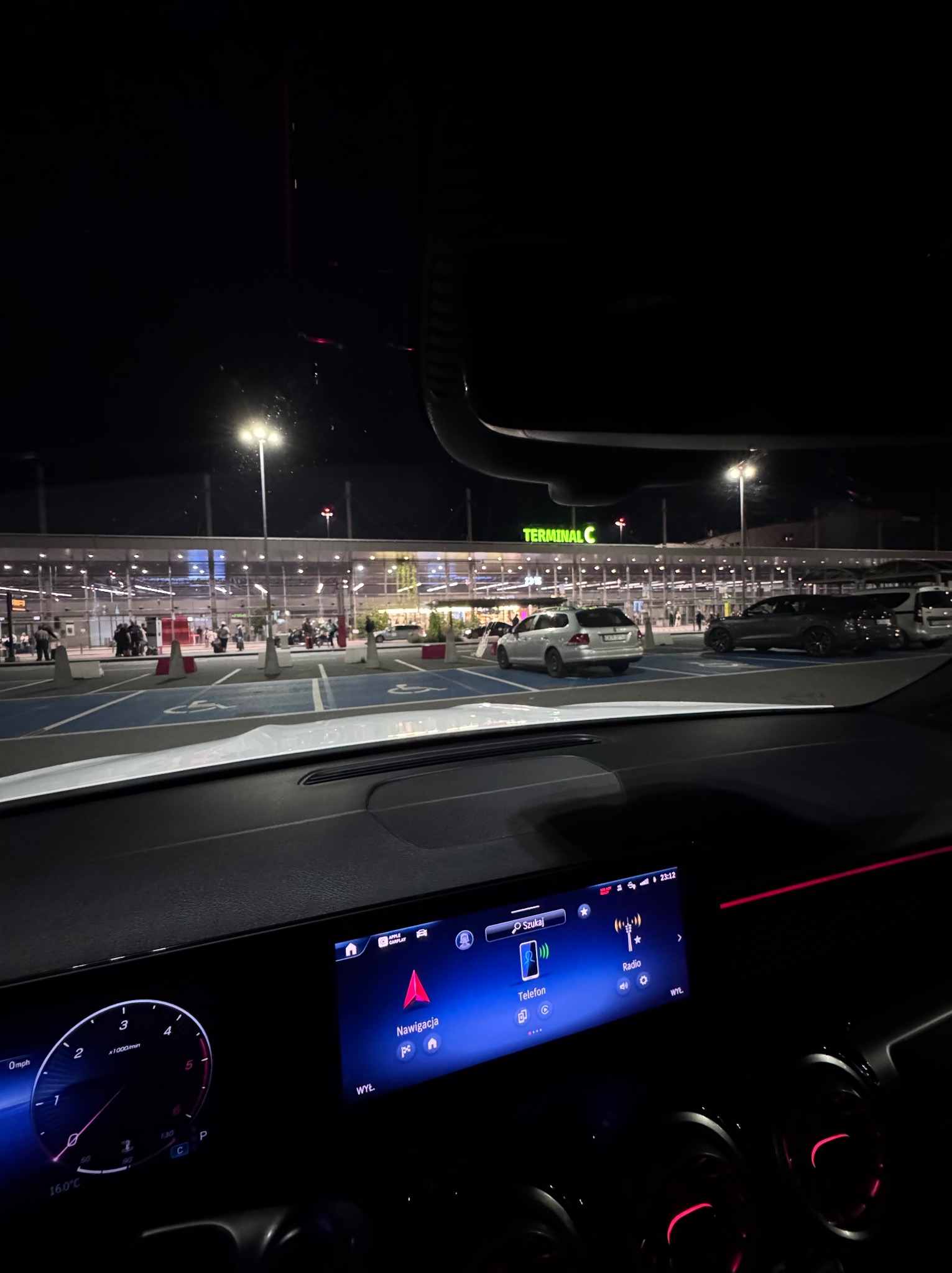 Widok z wnętrza samochodu na parking przed Terminalem C lotniska nocą. Na desce rozdzielczej włączony ekran nawigacji. Parkujące samochody i oświetlony budynek w tle.