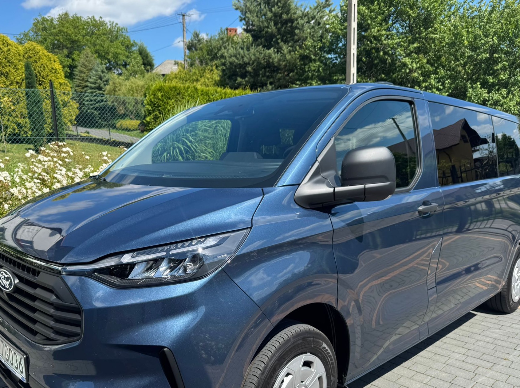 Niebieski Ford Transit Custom z reflektorem LED na tle zieleni i błękitnego nieba. Widoczny fragment tablicy rejestracyjnej.