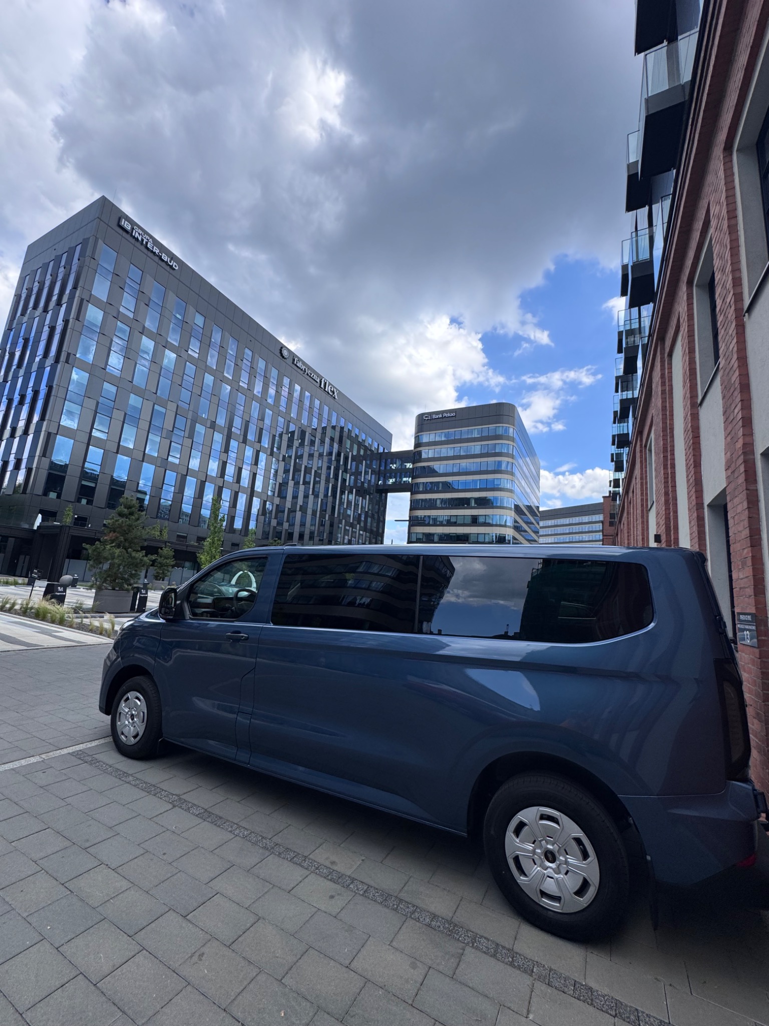 Niebieski bus na tle nowoczesnych biurowców z przeszklonymi fasadami i ceglanego budynku. Kadr z dołu, widoczne chmury.