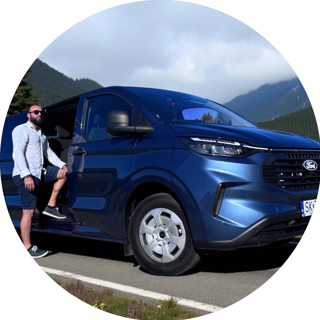 Niebieski Ford Transit z kierowcą na tle górskiego krajobrazu. Nowoczesny design, aluminiowe felgi i błękitny odcień szyby przedniej.