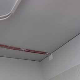STARPLAST MARCIN KOZAKOWSKI - Biały sufit po naprawie, widoczne rury w osłonie i fragment ściany z jasnymi płytkami. Na dole fragment samozamykacza do drzwi.