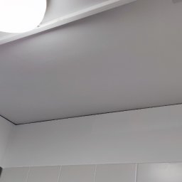 STARPLAST MARCIN KOZAKOWSKI - Biały sufit z zamontowanym oświetleniem, widoczne płytki na ścianie. Perspektywa z dołu, z bliska, ukazująca detale wykończenia i połączenia elementów.