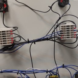 STARPLAST MARCIN KOZAKOWSKI - Wnętrze szafy sterowniczej z widocznymi elementami elektrycznymi, przewodami i złączkami, zabezpieczonymi opaskami. Widoczny fragment okna i rękaw skórzanej kurtki.