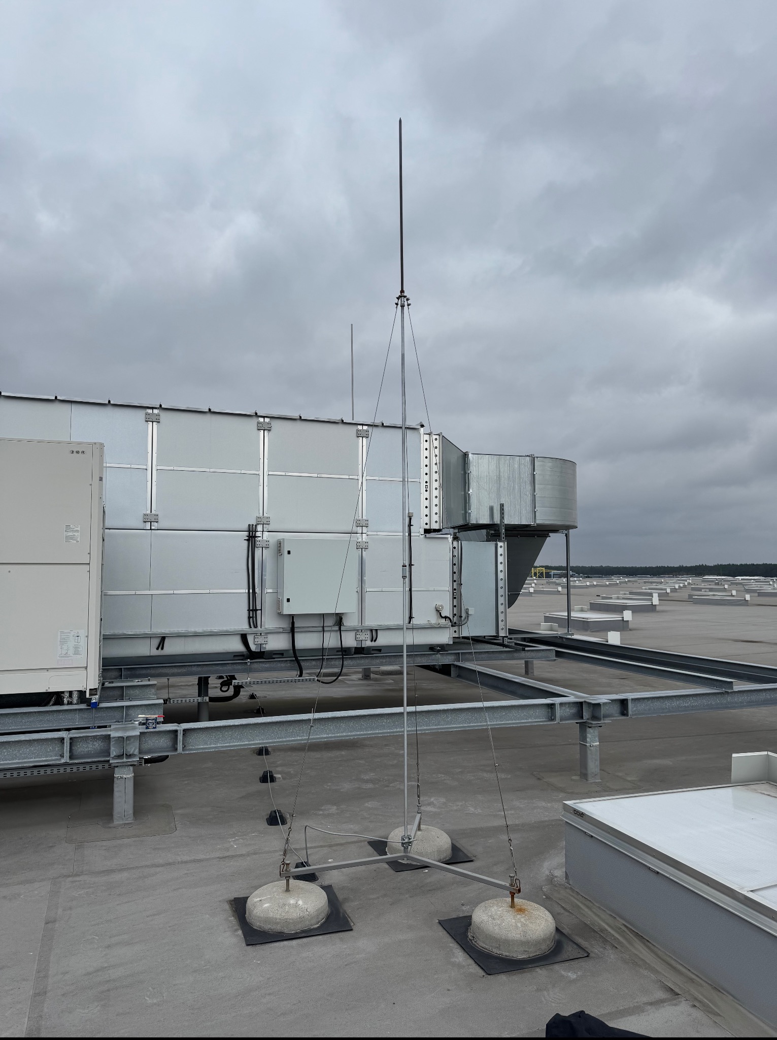 Maszt odgromowy z przewodami, zamocowany na betonowych podstawach na dachu hali magazynowej, widok z dołu na pochmurne niebo.
