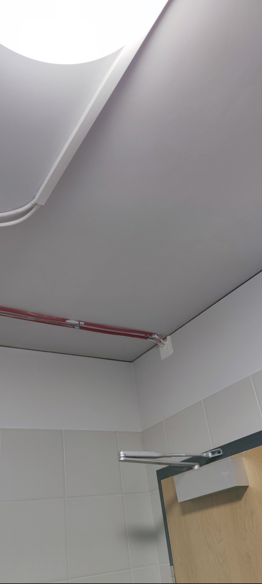 Biały sufit po naprawie, widoczne rury w osłonie i fragment ściany z jasnymi płytkami. Na dole fragment samozamykacza do drzwi.
