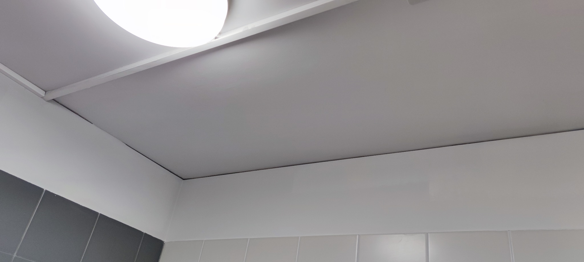 Biały sufit z zamontowanym oświetleniem, widoczne płytki na ścianie. Perspektywa z dołu, z bliska, ukazująca detale wykończenia i połączenia elementów.