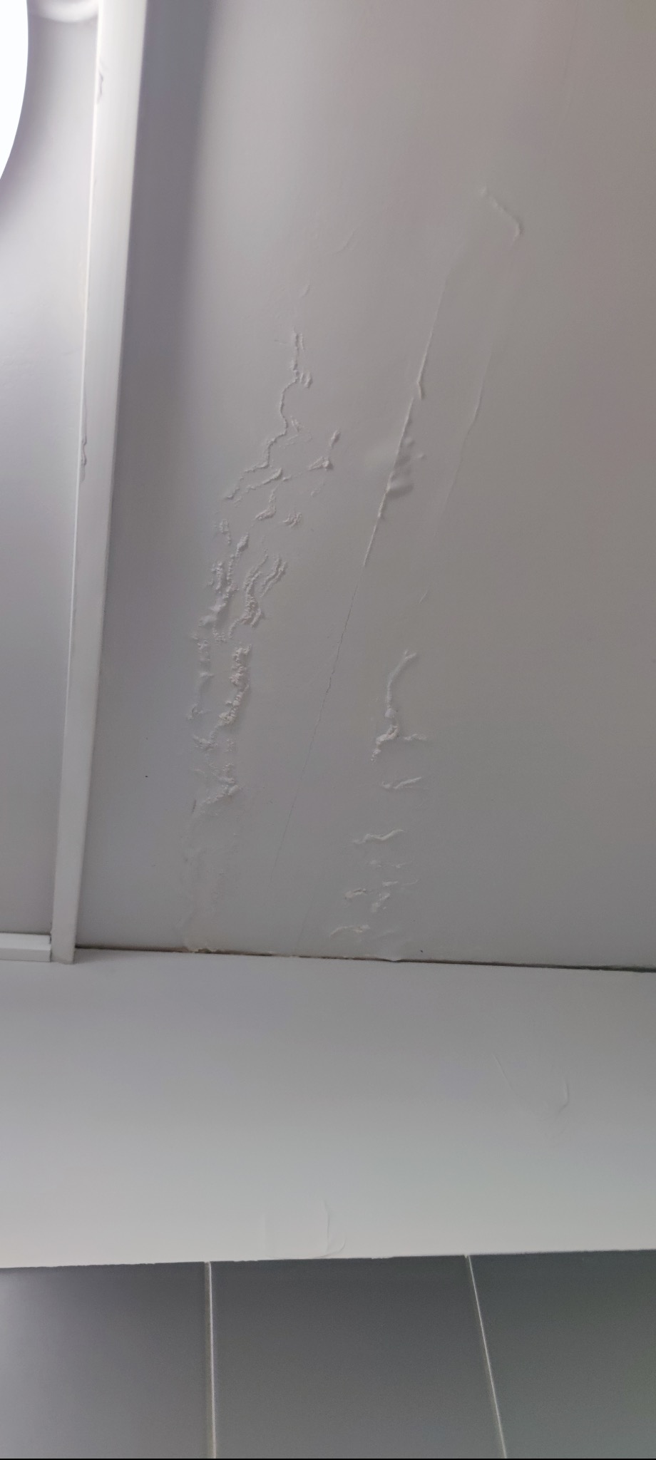 Z bliska widoczny biały sufit z wyraźnymi śladami zalania i uszkodzeniami. Widoczne wybrzuszenia i plamy świadczą o przecieku. Oświetlenie sufitowe w lewym górnym rogu.