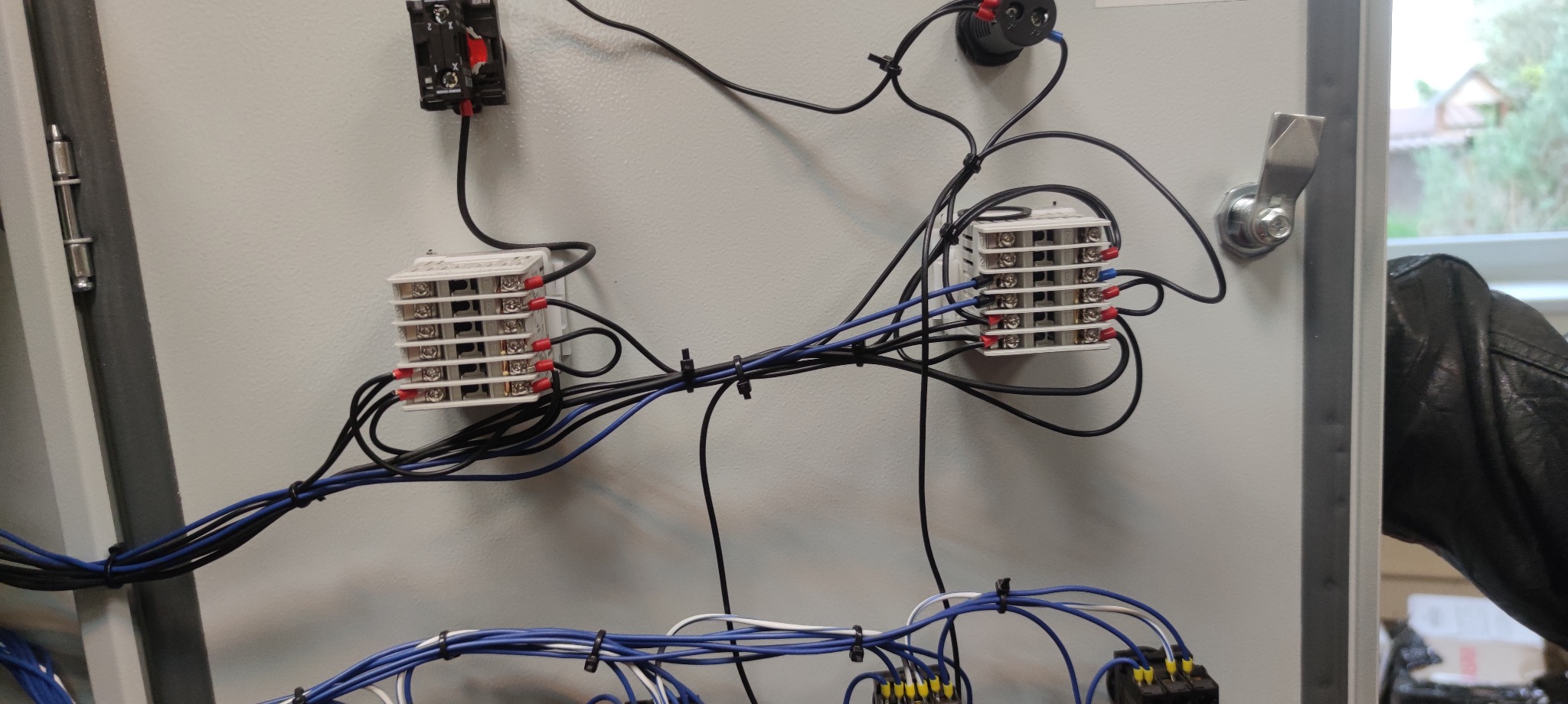 Wnętrze szafy sterowniczej z widocznymi elementami elektrycznymi, przewodami i złączkami, zabezpieczonymi opaskami. Widoczny fragment okna i rękaw skórzanej kurtki.