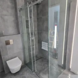 Szklana kabina prysznicowa z przesuwnymi drzwiami w szarej łazience z wiszącą toaletą i grzejnikiem drabinkowym. W tle widoczna drabina.