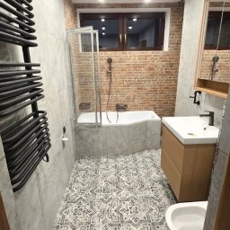 Sperbud - Wykończona łazienka z wanną, umywalką i grzejnikiem drabinkowym. Na podłodze wzorzyste płytki, ściany wyłożone szarymi płytkami imitującymi beton, a fragment ściany cegłą.