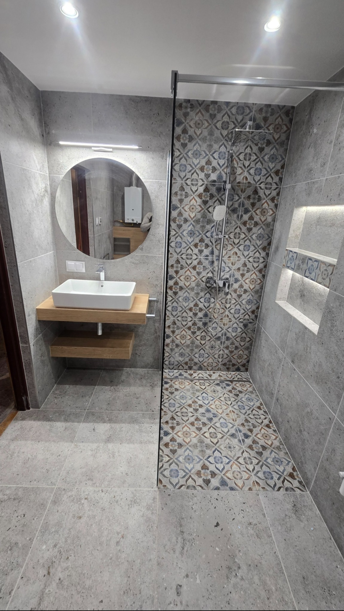 Nowoczesna łazienka z szarymi płytkami, okrągłym lustrem z podświetleniem, drewnianą szafką pod umywalką i kabiną prysznicową z wzorzystymi płytkami.