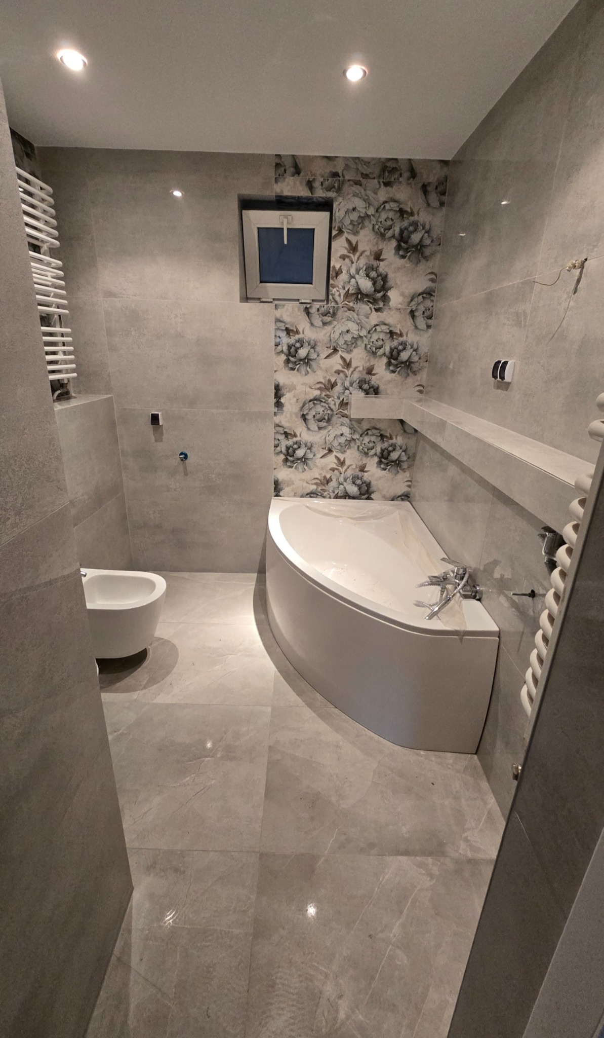 Wykończona łazienka z narożną wanną, bidetem i grzejnikiem drabinkowym. Szare płytki i ściana z motywem kwiatowym. Małe okno z niebieską szybą.