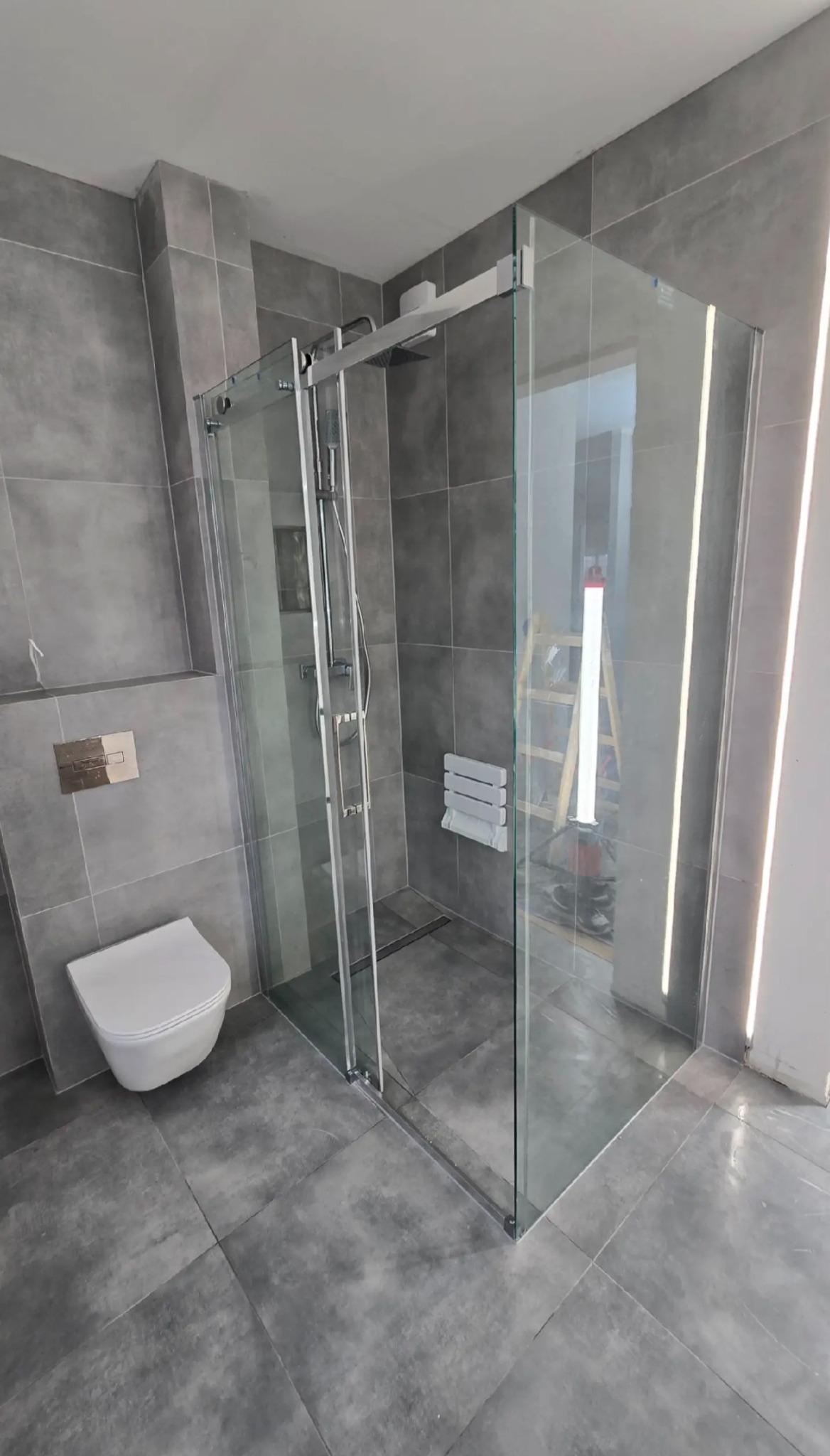 Szklana kabina prysznicowa z przesuwnymi drzwiami w szarej łazience z wiszącą toaletą i grzejnikiem drabinkowym. W tle widoczna drabina.