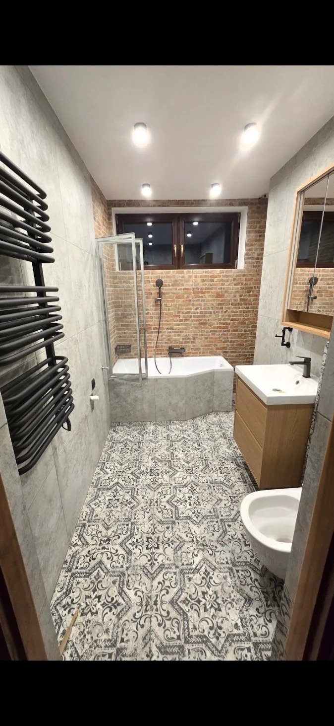 Wykończona łazienka z wanną, umywalką i grzejnikiem drabinkowym. Na podłodze wzorzyste płytki, ściany wyłożone szarymi płytkami imitującymi beton, a fragment ściany cegłą.