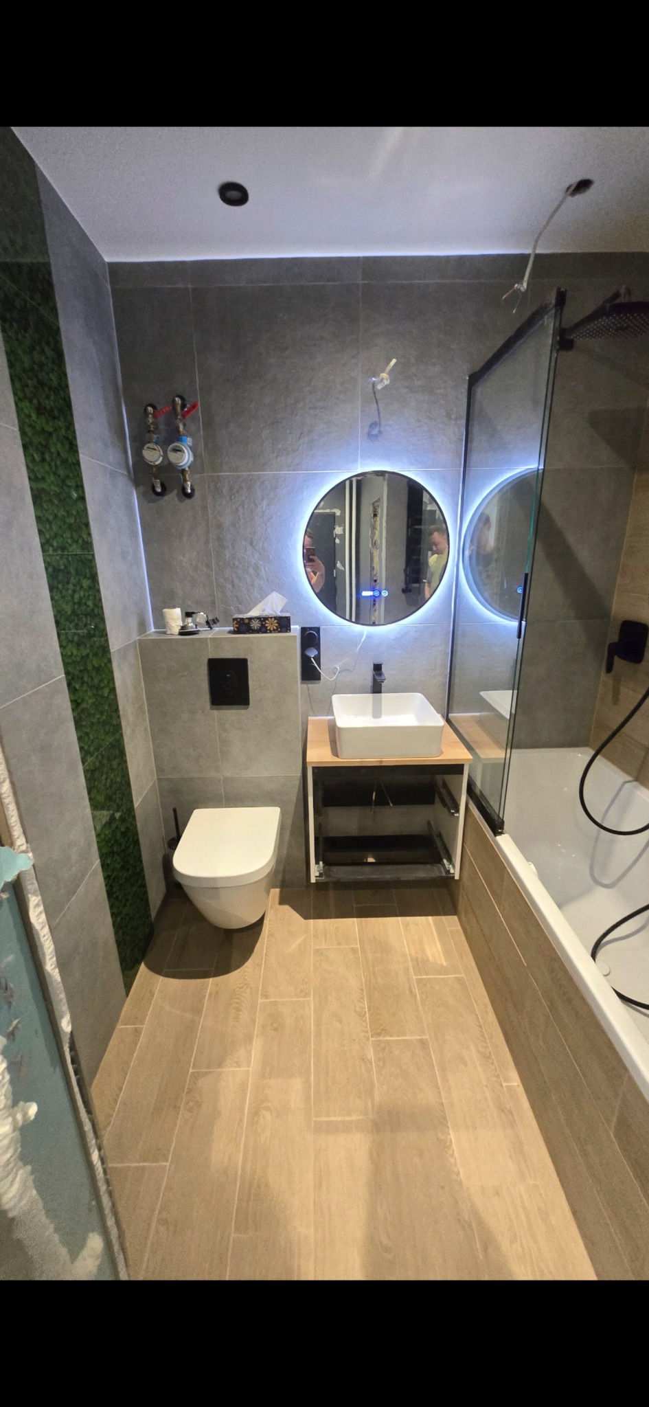 Nowoczesna łazienka po remoncie: szare płytki, imitacja zielonej ściany, podświetlane lustro, biała umywalka i toaleta. Jasne płytki podłogowe.