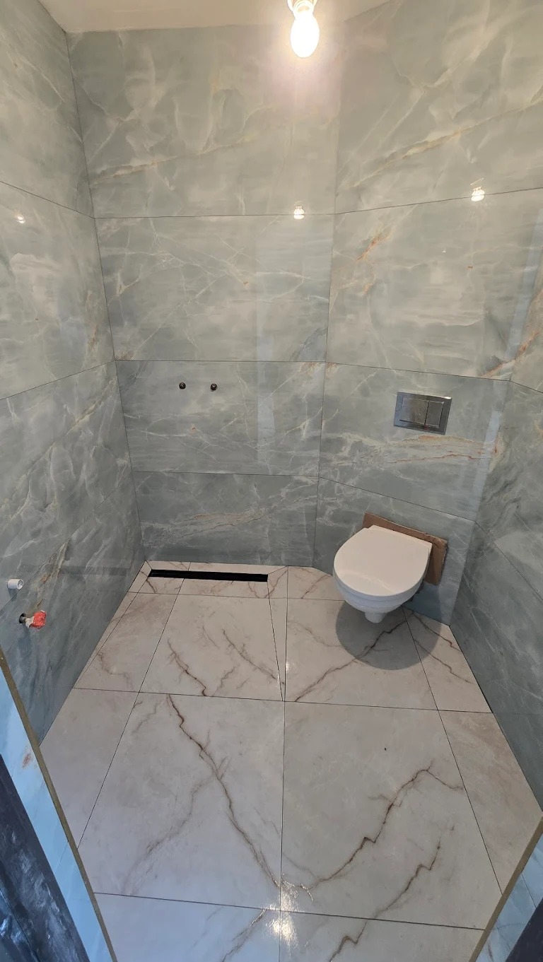 Wykończona łazienka z marmurowymi płytkami w odcieniach szarości i bieli. Widoczna toaleta, odpływ liniowy i armatura. Minimalistyczny design, jasne oświetlenie.