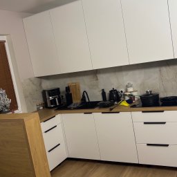 Prestigio - Nowoczesna kuchnia z białymi frontami i drewnianym blatem, czarne uchwyty, sprzęt AGD na blacie, marmurowy backsplash, oświetlenie punktowe.