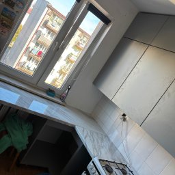 Prestigio - Szara kuchnia z białymi płytkami i widokiem na budynki przez okno. Ustawiona pod kątem, z kuchenką gazową i blatem imitującym marmur.