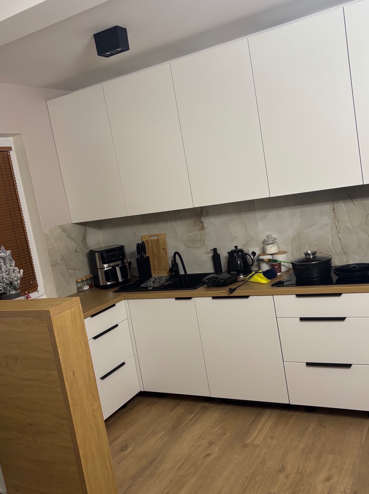 Nowoczesna kuchnia z białymi frontami i drewnianym blatem, czarne uchwyty, sprzęt AGD na blacie, marmurowy backsplash, oświetlenie punktowe.