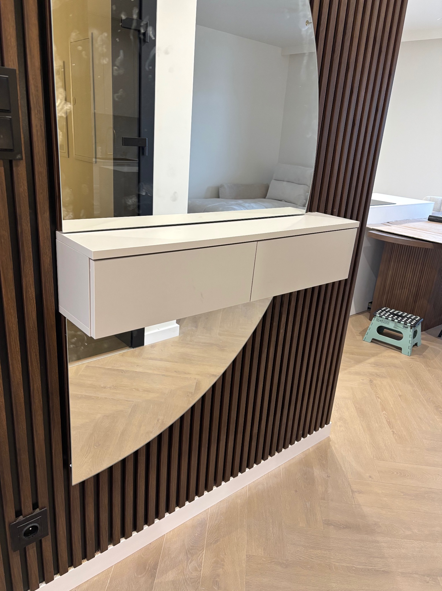 Nowoczesna szafka z lustrem w przedpokoju, z drewnianymi lamelami na ścianie i podłodze w jodełkę. Minimalistyczny design, jasne kolory, widoczne gniazdko elektryczne.