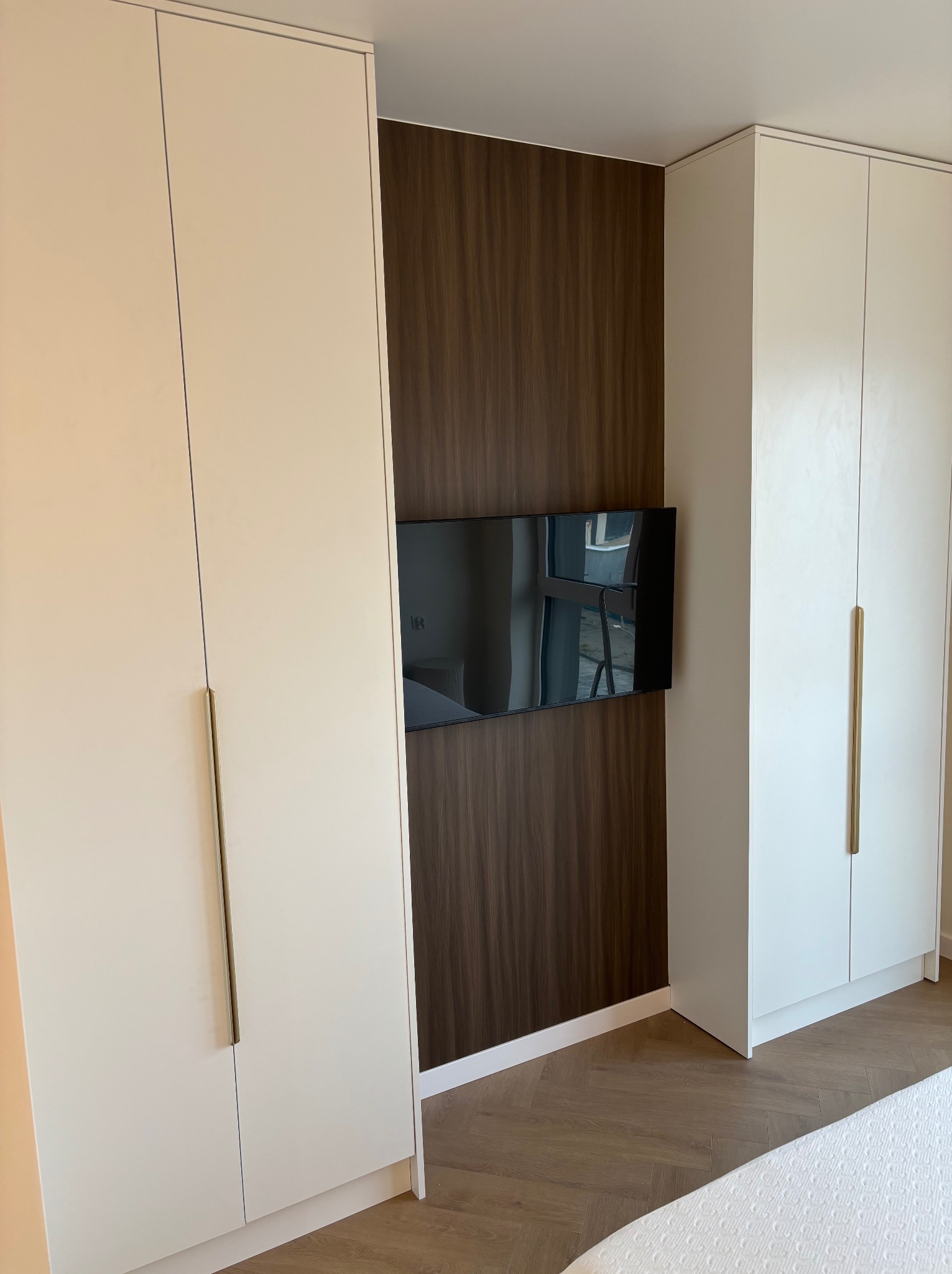 Białe szafy na wymiar z długimi, złotymi uchwytami, zamontowane na ścianie z drewnianą okładziną i telewizorem. Parkiet w jodełkę na podłodze.