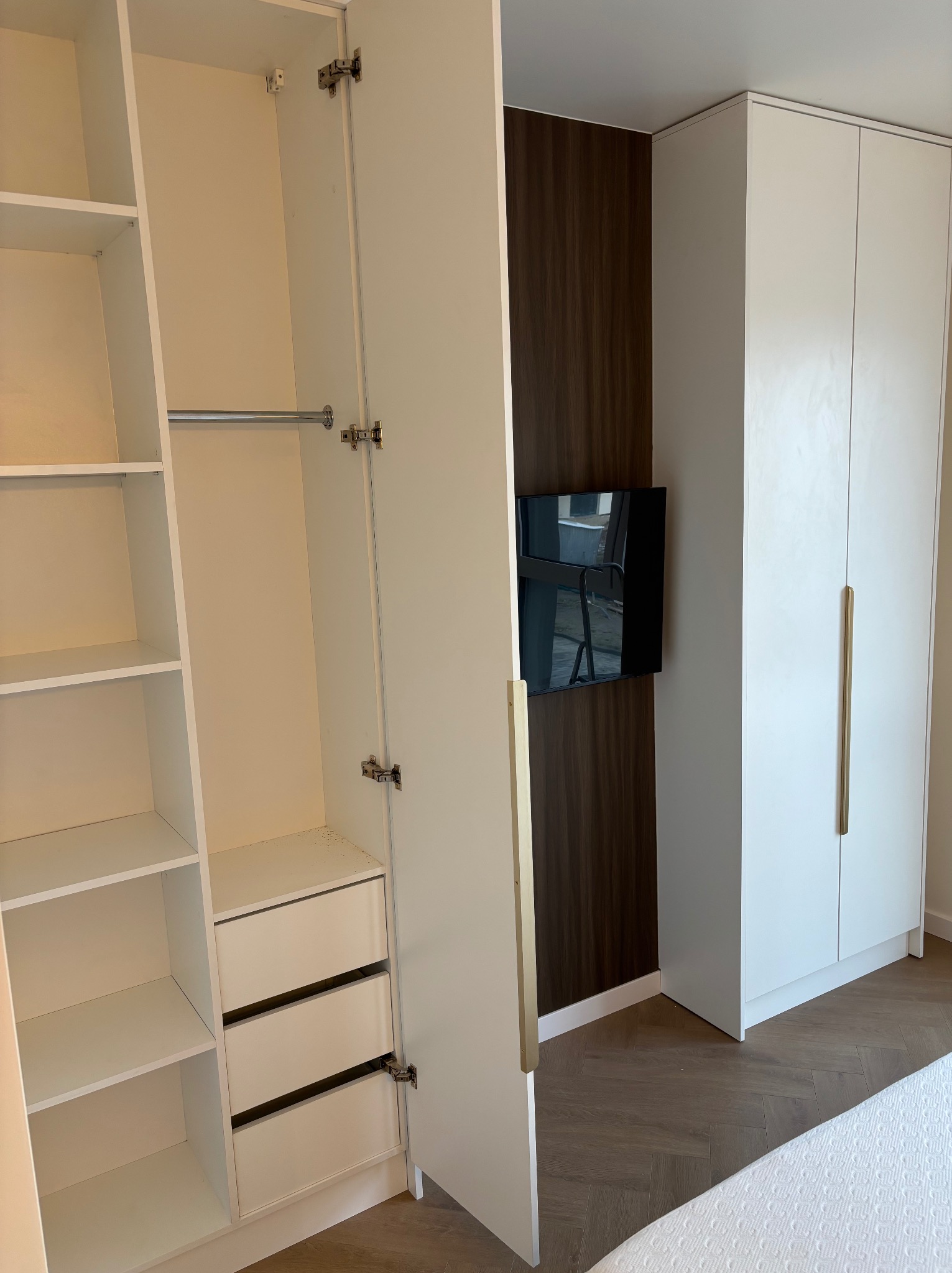 Biała szafa wnękowa z otwartymi drzwiami, widoczne półki i szuflady. Na ścianie obok telewizor i dekoracyjny panel. Podłoga z parkietu ułożonego w jodełkę.