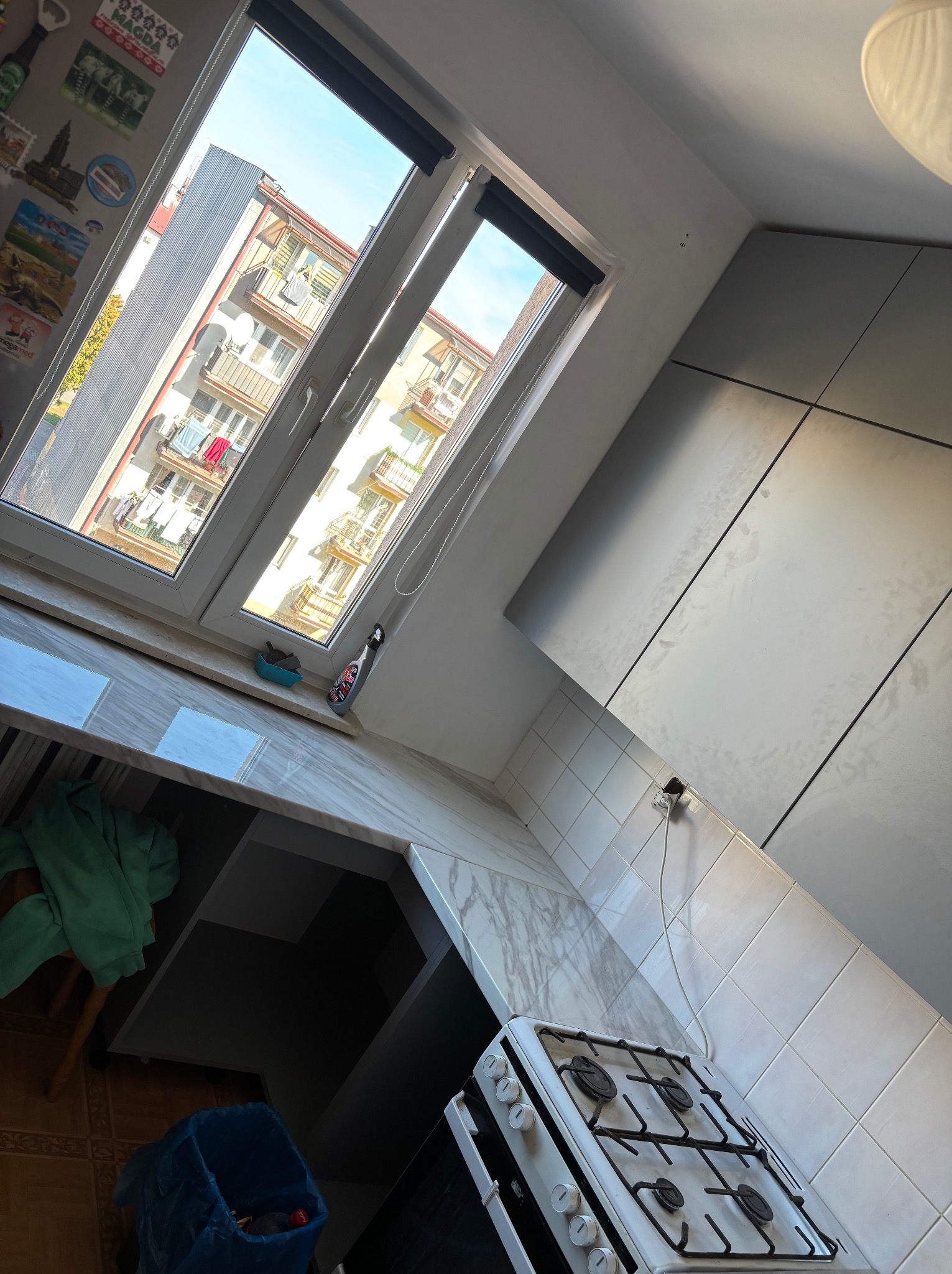 Szara kuchnia z białymi płytkami i widokiem na budynki przez okno. Ustawiona pod kątem, z kuchenką gazową i blatem imitującym marmur.