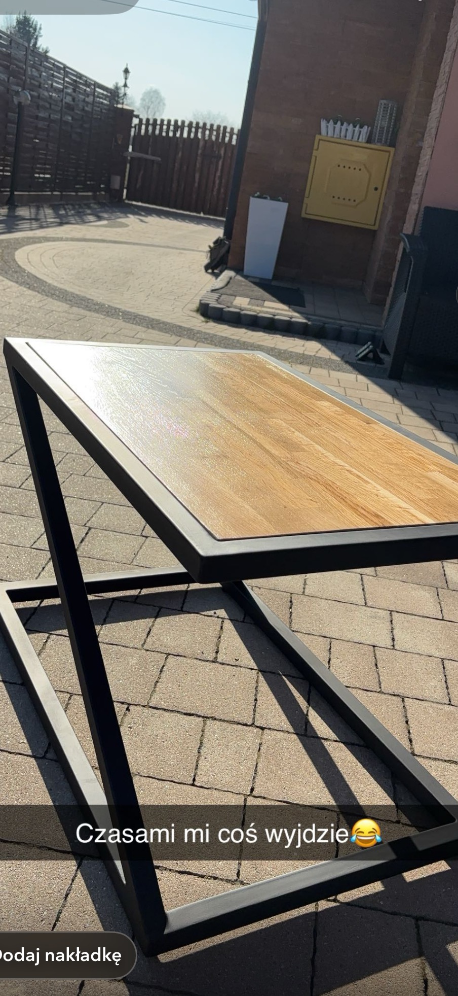 Stolik kawowy z drewnianym blatem i metalową, geometryczną podstawą w czarnym kolorze, ustawiony na brukowanym podwórku w słoneczny dzień.